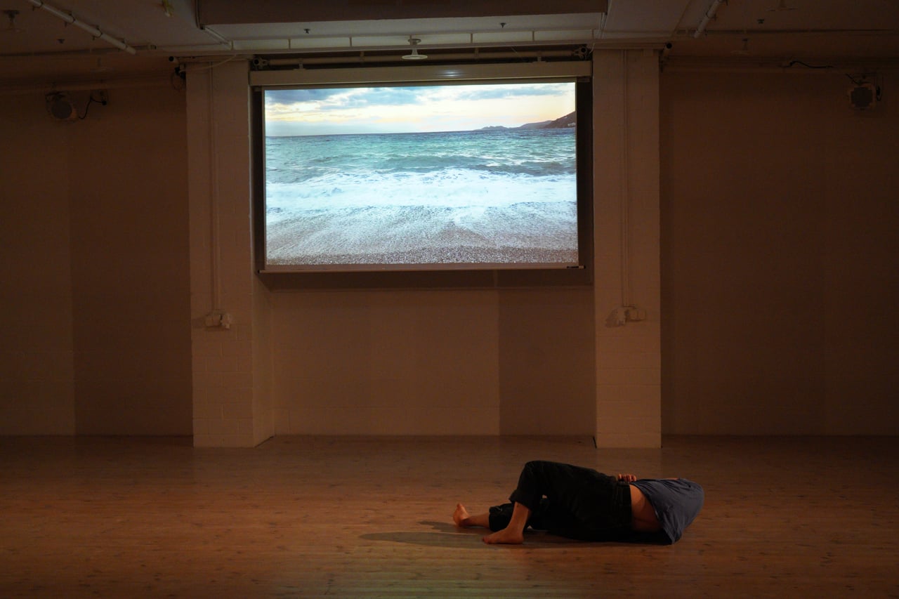 MOVING: a dancer lies on a wooden floor beneath a projection of the sea